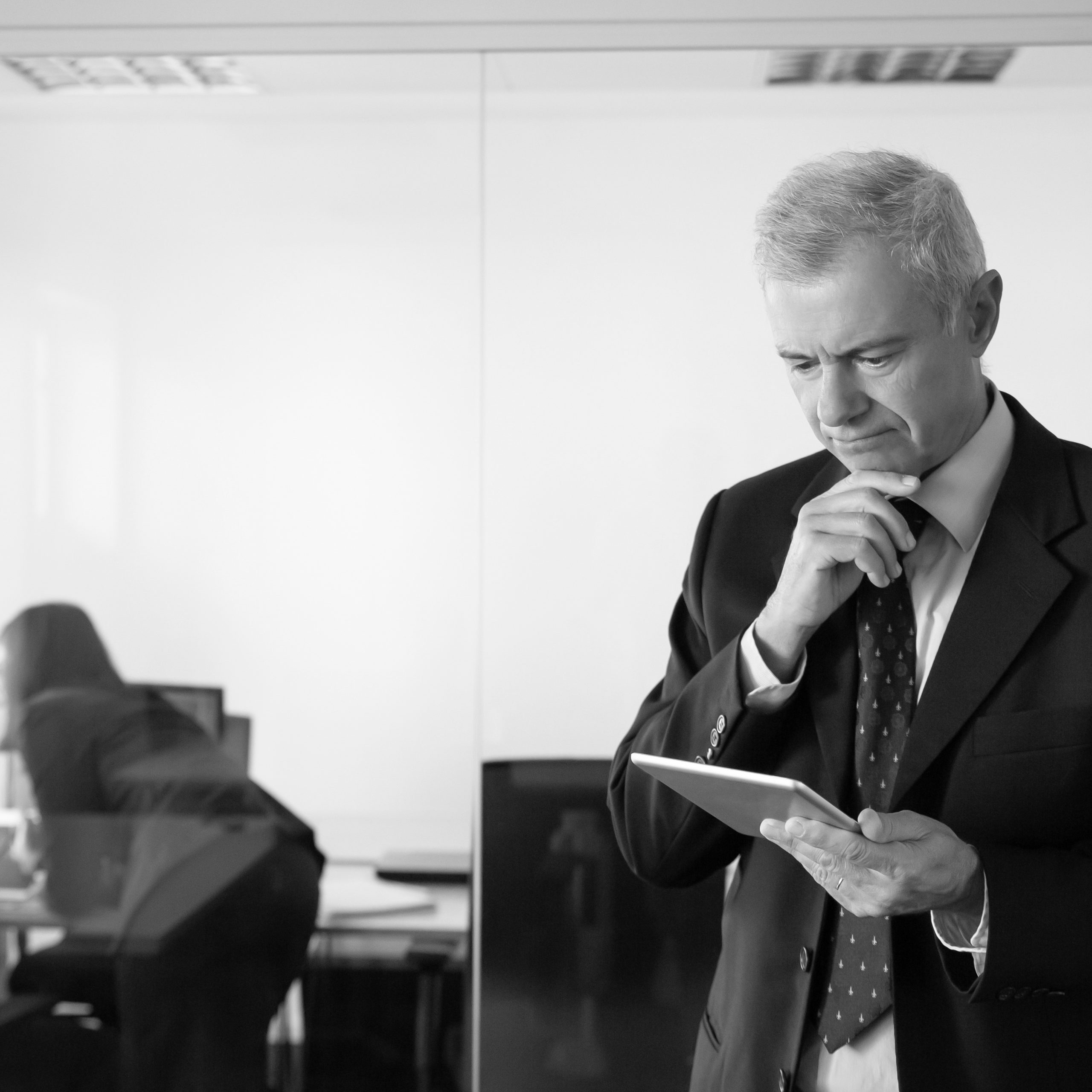 Focused pensive businessman staring at tablet screen while his colleagues discussing project at workplace behind glass wall. Copy space. Communication concept